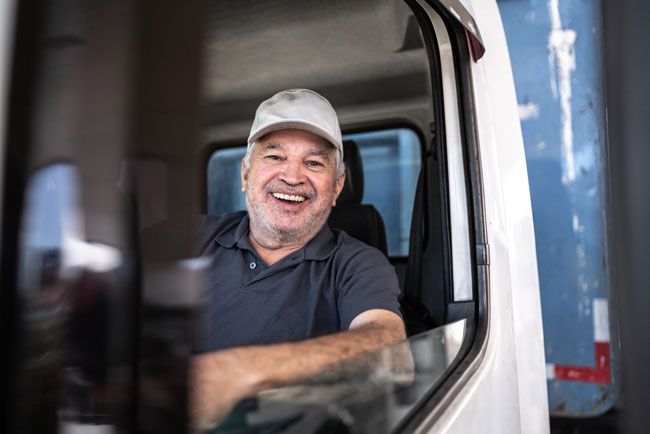 A fleet driver with gray hair wearing a ball cap is shown smiling out the window while he sits in the cab of his truck.