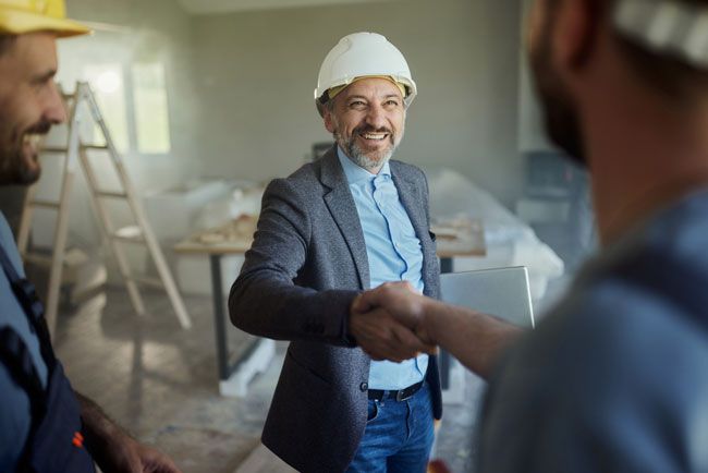 A construction company owner at a construction site is wearing a hard hat, holding a laptop with one hand and shaking the hand of his crew.