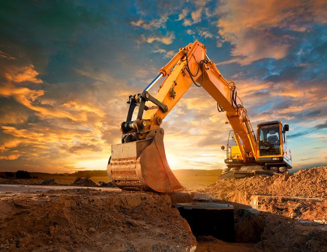 Image of a backhoe at a construction site with the sun setting in the background.